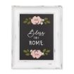 A framed decorative sign with a distressed white wooden frame and a black background. The sign features the text "Bless this home" in cursive and block letters, surrounded by pink and white flowers and green leaves at the top and bottom, created using the "transfer" by Chalk Couture.