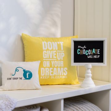 Show details for Maybe Chocolate Will Help Two decorative pillows are placed on a white bench. The larger yellow pillow reads, "DON'T GIVE UP ON YOUR DREAMS, KEEP SLEEPING." The smaller white pillow displays "DON'T DROP THE f" with a cartoon bomb illustration. A letterboard next to them says, "Maybe CHOCOLATE WILL HELP?". All the designs were created using the Chalk Couture transfer.