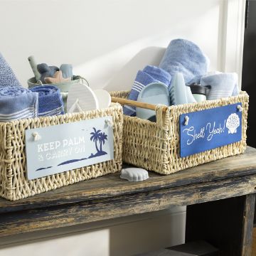 Show details for Shell Yeah! Two woven baskets sit on a rustic wooden shelf. One basket's sign reads "KEEP PALM & CARRY ON" with a palm tree, and the other reads "Shell Yeah!" with a seashell. Both are decorated using the transfer from Chalk Couture and filled with blue and white bath items like towels, flip-flops, and brushes.