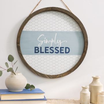 Show details for Blessed Simply A round wall decoration with a wood frame and chicken wire backing reads "Simply Blessed" in white and blue text. Below the sign, a Chalk Couture transfer, a white vase with eucalyptus leaves, a blue book, and two small cream-colored jars are arranged on a light-colored table.