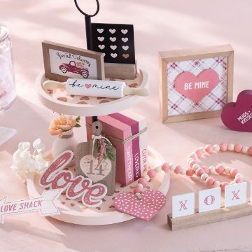 Show details for Loveshack A decorated display for Valentine's Day featuring a two-tiered tray holding small pink and red items, including a "love" sign, a heart-shaped decor, and Valentine's cards. Surrounding items include a "Be Mine" framed sign, a bead garland, and more Valentine's-themed decor.