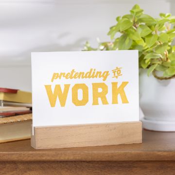 Show details for Pretending to Work A white sign with the text "pretending to WORK" written in bold yellow letters is displayed in a wooden holder on a desk. In the background, there is a green plant in a white pot and some office supplies partially visible. The sign was created using the transfer from Chalk Couture.