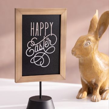 Show details for Happy Easter Swirls A small blackboard with a wooden frame reads "Happy Easter" in white cursive text. The board is standing on a black base. Next to it is a decorative rabbit statue made of a light brown material with intricate carving details. The background is blurred. This beautiful display was created using the "Happy Easter" transfer from Chalk Couture.