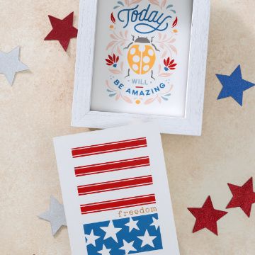 Show details for Today Will Be Amazing, Freedom A framed artwork with a ladybug and the text "Today will be amazing" sits next to a Chalk Couture transfer featuring a stylized American flag with red stripes and blue stars, including the word "freedom." Red and silver star-shaped decorations surround the items.