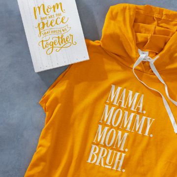Show details for Mama Mommy Mom A yellow hoodie with white text that reads "MAMA. MOMMY. MOM. BRUH." is displayed against a gray background. Next to it is a white wooden sign with gold text that says "Mom, you are the piece that holds us together," created using a Chalk Couture transfer.