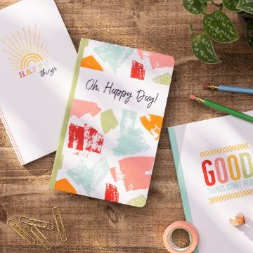 Show details for Good Things Start Here A wooden table holds colorful stationery items, including a Chalk Couture transfer with a cover that reads "Oh, Happy Day!" in a vibrant design, another Chalk Couture transfer with "HAPPY things" and a sun illustration, and a third with "GOOD things start here." Nearby are pencils, washi tape, and paper clips.