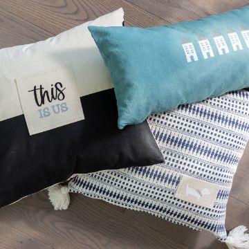 Show details for This Is Us Three decorative pillows on a wooden floor. The first pillow is black and cream with a "this is us" patch created using the transfer from Chalk Couture. The second pillow is teal with a pattern of houses. The third pillow features a blue and white geometric design with a small patch of a sheep made using the transfer from Chalk Couture.