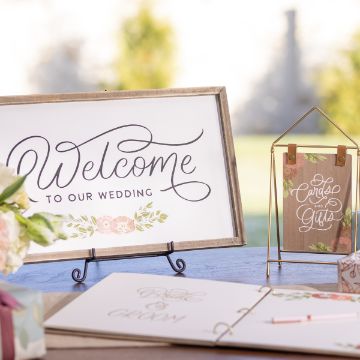 Show details for Welcome to Our Wedding A wedding welcome sign with elegant calligraphy reading "Welcome to our wedding" is displayed on a stand. Next to it, a card holder labeled "Cards and Gifts" sits on a table decorated with flowers and a guestbook. The background is out of focus.