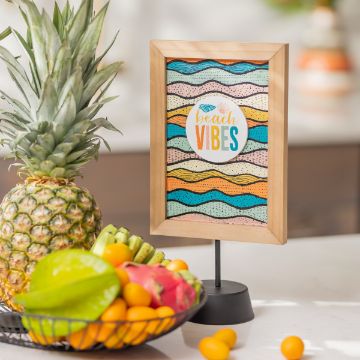 Show details for Geometric Ocean Prints A framed artwork with the text "beach vibes" stands on a table. The frame features a colorful, wavy pattern. In front, there's a bowl of assorted tropical fruits including bananas, oranges, a green apple, and a pineapple. The background is blurred.