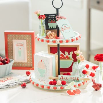 Show details for Farmhouse Cherries A decorative two-tier tray holds small books and a variety of charming items, including flowers, cherries, and signs reading "Sweet Cherries" and "Grandma's Kitchen." Other related decor, such as framed pictures and more Chalk Couture transfer designs, are arranged around the tray.