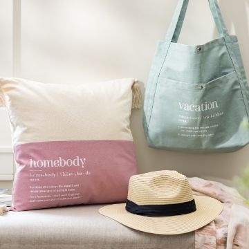 Show details for Vacation Homebody A beige and pink cushion with the word "homebody" and its definition printed on it sits on a light-colored sofa. Next to it, a light green tote bag with the word "vacation" and its definition hangs over the sofa. A Chalk Couture transfer rests nearby.