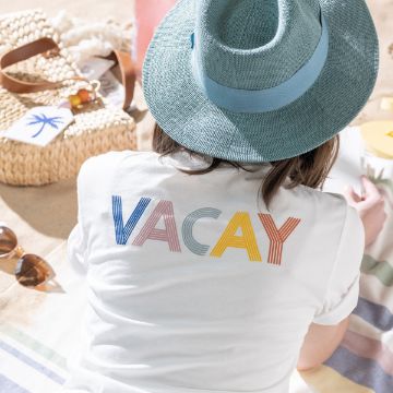 Show details for Vacay Mode A person wearing a blue hat and a white t-shirt with the word "VACAY" in multicolored letters lies on a beach blanket. Nearby are sunglasses, a woven bag with a palm tree design, and other Chalk Couture transfer beach accessories. Sand is visible, indicating they are on a beach.