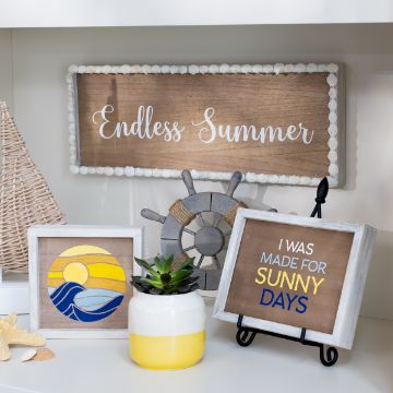 Show details for Sunkissed A shelf displays a coastal-themed decor arrangement. A wooden sign reads "Endless Summer." Below it, a framed artwork of the sun and waves sits next to a small yellow and white pot with a plant. Another framed sign reads "I was made for sunny days," which features the transfer from Chalk Couture.