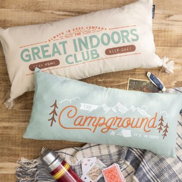 Show details for Always in Good Company Two rectangular throw pillows are placed on a wooden surface. The top pillow reads "Great Indoors Club: Stay Home, Keep Cozy" while the bottom one says "The Campground: It's 'in tents'." There is also a gray plaid blanket, a deck of cards, and a red thermos beside the pillows.