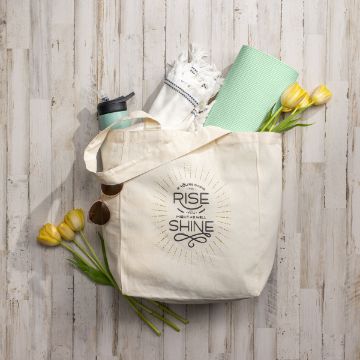 Show details for You Might As Well Shine A canvas tote bag with the phrase "You’re Going to Rise and Might as Well Shine" printed on it. The bag contains a green yoga mat, a white fringed towel, a light blue water bottle, and has yellow tulips and sunglasses draped over it.