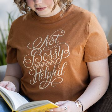 Show details for Not Bossy A person wearing a brown t-shirt with the text "Not Bossy, Aggressively Helpful" written in white cursive script is sitting and reading a book. The person has curly hair and is indoors with a plant visible in the background. The design on the t-shirt was created using the transfer from Chalk Couture.