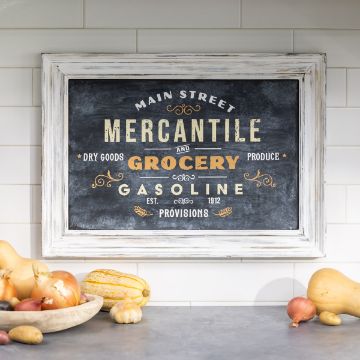 Show details for Main Street Mercantile A framed chalkboard sign with vintage-style text advertising "Main Street Mercantile and Grocery," "Dry Goods," "Produce," and "Gasoline, Est. 1912." Various vegetables, including onions and squash, are placed on a countertop below the sign. This design was created using a transfer from Chalk Couture.