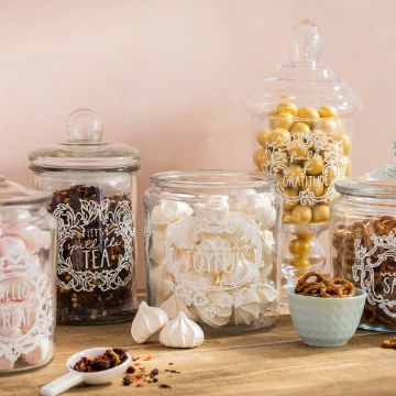 Show details for Crockery Labels A collection of glass jars with ornate white designs, each containing different items. The jars hold dried tea, pastel-colored meringues, gold-coated candies, and pretzels. A small blue bowl with pretzels and a white spoon with tea leaves are also on the wooden surface, decorated using a Chalk Couture transfer.