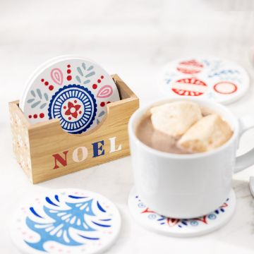 Show details for Coaster Set (7 Pieces) A wooden box with the word "NOEL" holds festive, decorated coasters featuring red and blue designs. One coaster is in use under a white mug filled with hot chocolate topped with marshmallows, and other coasters are scattered nearby on a white surface.