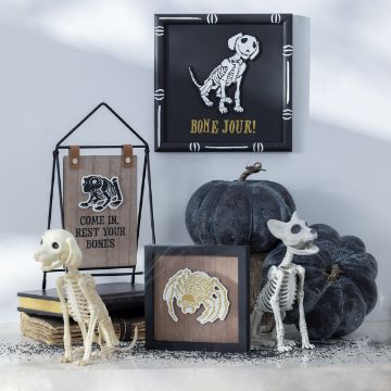 Show details for Couture Boutique® Chalkable Shapes Bad to The Bone (5 Pieces) A Halloween-themed display featuring framed skeleton art, blue pumpkins, and decorative skeleton figures of a dog and a cat. Signs include phrases "BONE JOUR!" and "COME IN, REST YOUR BONES" against a neutral background.