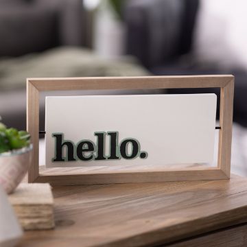 Show details for Hello, Goodbye Reusable Shapes (2 Pieces) A wooden frame with a white panel inside displays the word "hello" in bold, dark green lowercase letters. The frame is sitting on a wooden surface, with a blurred background that includes a couch and some greenery.