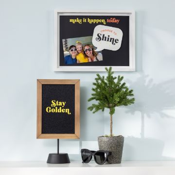 Show details for Couture Boutique® Chalkable Shapes You Are Awesome A tabletop scene features a small potted tree, black-framed sunglasses, and a sign with the yellow text "Stay Golden." Above is a framed collage with a photo of three people and the phrases "make it happen, today" and "CHOOSE TO Shine".