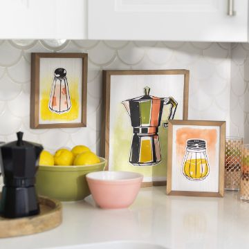 Show details for Salt & Pepper A kitchen counter adorned with framed artwork of kitchen items, including a salt shaker, stovetop coffee maker, and a mason jar. There are fresh lemons in a green bowl, a black stovetop coffee maker, a pink bowl, and a small mosaic-patterned glass. The artwork features the Chalk Couture transfer for added detail.