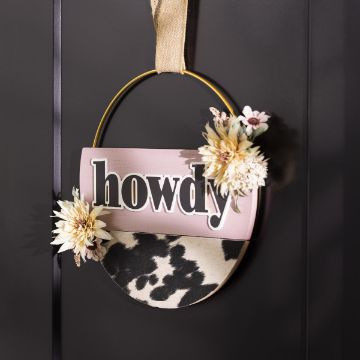 Show details for Welcome Words A decorative sign hangs on a black wall, featuring the word "howdy" in bold letters. The sign is adorned with faux flowers and has a cowhide-patterned lower section. It hangs from a beige ribbon attached to a circular frame, all designed with the Chalk Couture transfer.