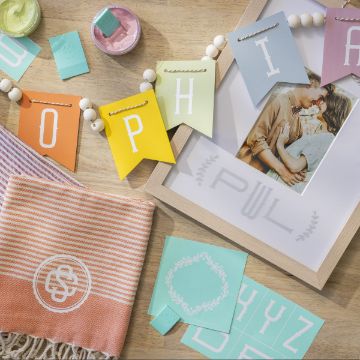 Show details for Modern Monogram Font A wooden table displays a colorful "SOPHIA" banner, pastel art supplies, fabric, and a framed photo of a couple embracing. Mint green, pink, and yellow items include letters and decals, adding a crafty, personalized touch to the setting.