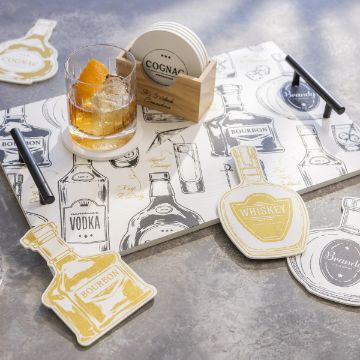 Show details for But First Cocktails Reusable Shapes (6 Pieces) A table with coasters and napkins featuring drawings of various liquor bottles, including bourbon, vodka, and whiskey. A glass of whiskey with ice and an orange peel garnish sits on a coaster. A set of coasters is held in a wooden holder labeled "COGNAC.
