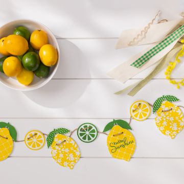 Show details for Lemons A white table displays a bowl of lemons and limes on the left, with a bundle of ribbons in various colors and textures on the top right. Below, a decorative garland featuring lemons, limes, and the phrase "Hello Sunshine" is strung across the table.
