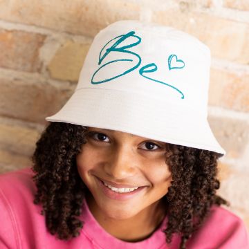 Show details for Be Salty A smiling person with curly hair wears a white bucket hat featuring teal text that says "Be" and includes a heart design. The person is dressed in a pink top and stands in front of a light brick wall, showcasing the transfer from Chalk Couture.