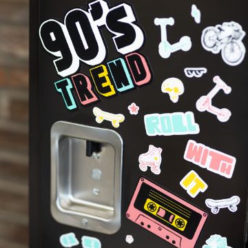Show details for Roll With It Retro A black surface is decorated with an assortment of colorful stickers, featuring text like "90’s Trend" and "Roll With It," along with images of a cassette tape, roller skates, scooters, bikes, a skateboard, and various small icons. There's also a metal rectangular fixture. This design is created using the transfer from Chalk Couture.