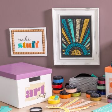 Show details for Make Stuff A crafting corner with a purple wall displays two framed artworks: one with "make stuff" text and another with a colorful geometric sunburst design. A white box labeled "art stuff", paint jars, and Chalk Couture transfer crafting supplies are organized on the table.