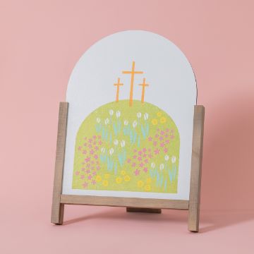 Show details for Hill & Crosses A Chalk Couture transfer on a wooden stand shows three crosses on a hilltop. The hill is adorned with white tulips, purple flowers, and yellow flowers. The background is a solid light pink color.