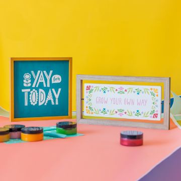 Show details for Grow Your Own Way A colorful crafting setup featuring two framed quotes. The left frame has a teal background with "YAY for TODAY" in white, playful text. The right frame has a floral border around the words "GROW YOUR OWN WAY". Various Chalk Couture transfer paint containers are visible in the foreground.
