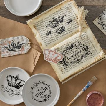 Show details for Reusable Vintage Labels & Crowns Shapes (3 Pieces) A vintage-themed flat lay featuring an open book with printed crown illustrations, decorative tags, a paintbrush, and plates with French patisserie designs. The items are placed on a brown paper background, evoking an antique, artisanal aesthetic.