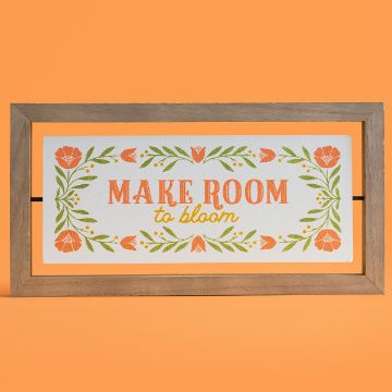 Show details for Make Room to Bloom A rectangular wooden-framed sign with a white background displays the text "MAKE ROOM to bloom" in orange and yellow. The sign is adorned with floral and leafy motifs in orange and green, set against an orange backdrop. This artistic creation was achieved using the transfer from Chalk Couture.