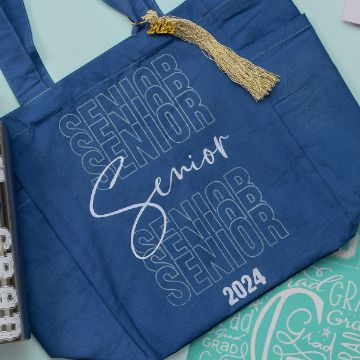 Show details for Senior A blue tote bag with the word “Senior” repeatedly printed in white. In the foreground, "Senior" is written in a cursive script and "2024" is printed at the bottom. The bag also features a graduation tassel. Other Chalk Couture transfer items are partially visible.