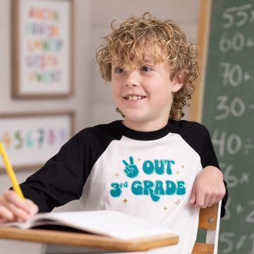 Show details for Peace Out A young child with curly hair wearing a white and black shirt that reads "Peace Out 3rd Grade" is sitting at a desk, smiling while holding a pencil. A Chalk Couture transfer is in the background, along with colorful numbers and letters on the wall.