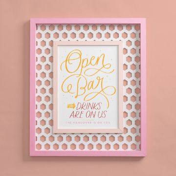 Show details for Open Bar Framed artwork with the caption "Open Bar. Drinks are on us. The hangover is on you." in yellow and pink cursive text made using Chalk Couture transfer. The frame is pink with a white honeycomb-patterned border, set against a light pink wall.