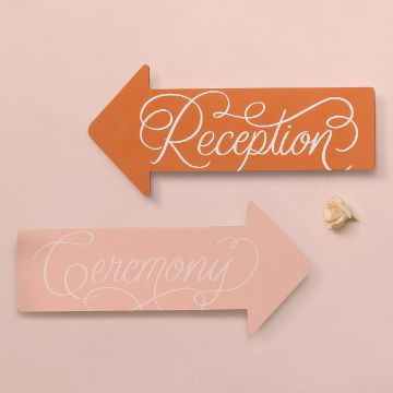 Show details for Reception Two directional arrow signs on a light pink background. The upper orange sign points left and reads "Reception" in elegant script. The lower light pink sign points right and reads "Ceremony" in similar elegant script. A small white rose is placed nearby.