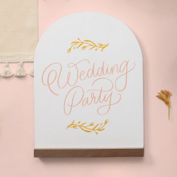 Show details for Wedding Party A white arched sign with the words "Wedding Party" written in pink cursive. Yellow decorative leaves embellish the sign. A brown shelf is attached at the bottom. The background is a light pink surface with a small orange flower and a fringed ivory fabric.