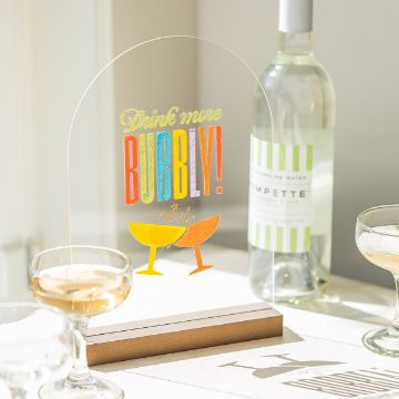 Show details for Drink More Bubbly A festive scene with a sign reading "Drink more Bubbly!" Two champagne glasses are depicted clinking under the sign. A bottle of white wine and filled champagne glasses are on the table, capturing a celebratory atmosphere with sunlight streaming in. The sign was created using a transfer from Chalk Couture.