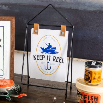 Show details for Keep It Reel A Chalk Couture transfer with a fishing theme is displayed on a black wire stand. The transfer reads "Gone Fishing, Be Back Soon, Keep It Reel" with an illustration of a person fishing in a boat and an anchor. Various fishing-related items surround the transfer on the wooden surface.