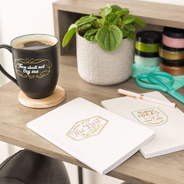 Show details for Brilliant Ideas A wooden desk holds a black coffee mug with a decorative quote, three white notebooks with colorful labels, a green potted plant in a white textured pot, and stationery items including scissors and tape. The scene is well-lit and organized, featuring the Chalk Couture transfer.