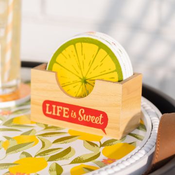 Show details for Sweet Citrus A wooden stand with the words "LIFE is Sweet" holds round coasters designed like slices of lemon. The stand is placed on a table covered with a floral-patterned tablecloth. A glass with a striped straw is partially visible on the left, created using the transfer from Chalk Couture.