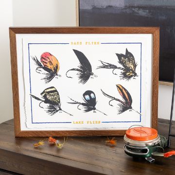 Show details for Lake Flies Framed Chalk Couture transfer showing six illustrated fishing flies labeled as “Bass Flies” and “Lake Flies.” Each fly is individually numbered and intricately detailed. The frame is placed on a wooden surface next to a fishing reel and tackle.