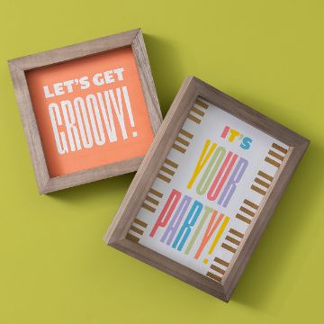 Show details for Happy Day Two framed signs on a yellow surface. The left sign has an orange background with the Chalk Couture transfer text "LET'S GET GROOVY!" in white and yellow. The right sign has a white background with colorful Chalk Couture transfer text "IT'S YOUR PARTY!" bordered by vertical gold lines.