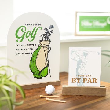 Show details for Best Dad By Par A desk display features two golf-themed signs. The larger one reads "A bad day of golf is still better than a good day at work" with an illustration of a golf bag. The smaller sign shows a golfer in mid-swing with the text "Best Dad by Par." A Chalk Couture transfer and tees are in the foreground.